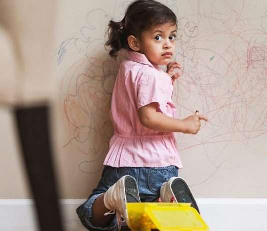 دروغ گویی در کودکان، کودکی مشغول نقاشی روی دیوار است و لباس صورتی رنگ و شلوار جین پوشیده و نگران در حال نگاه به پشت سرش است